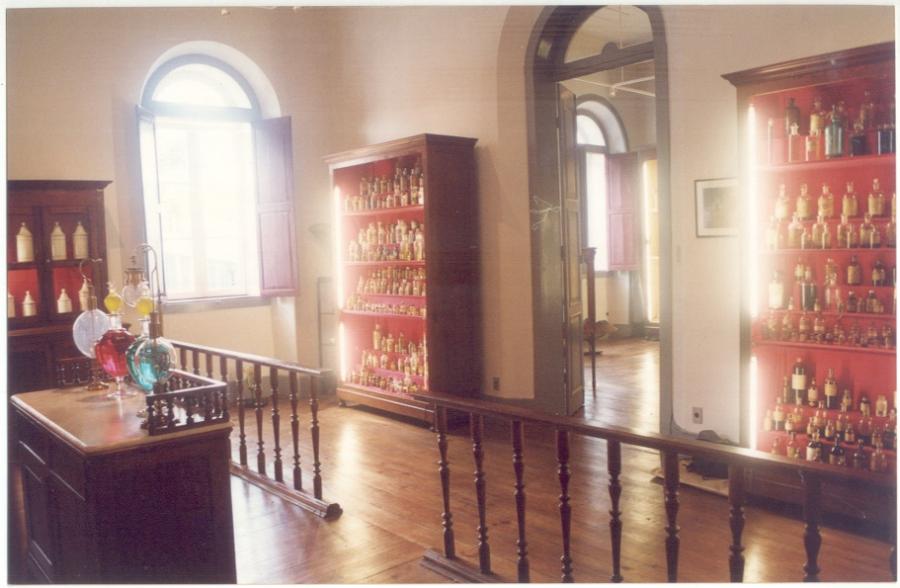 Posicionamento da Farmácia Magalhães Gomes com extratos, tinturas e vidrarias na 1ª sala e sua reorganização na 2ª sala.